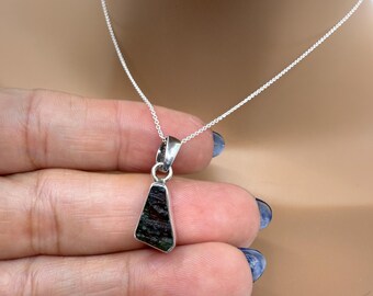 May include: A silver necklace with a pendant featuring a dark green, triangular stone. The stone has a speckled pattern. The necklace is displayed against a neutral background.