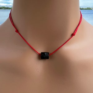 Puede incluir: Un collar de cuerda roja con un colgante cuadrado negro.