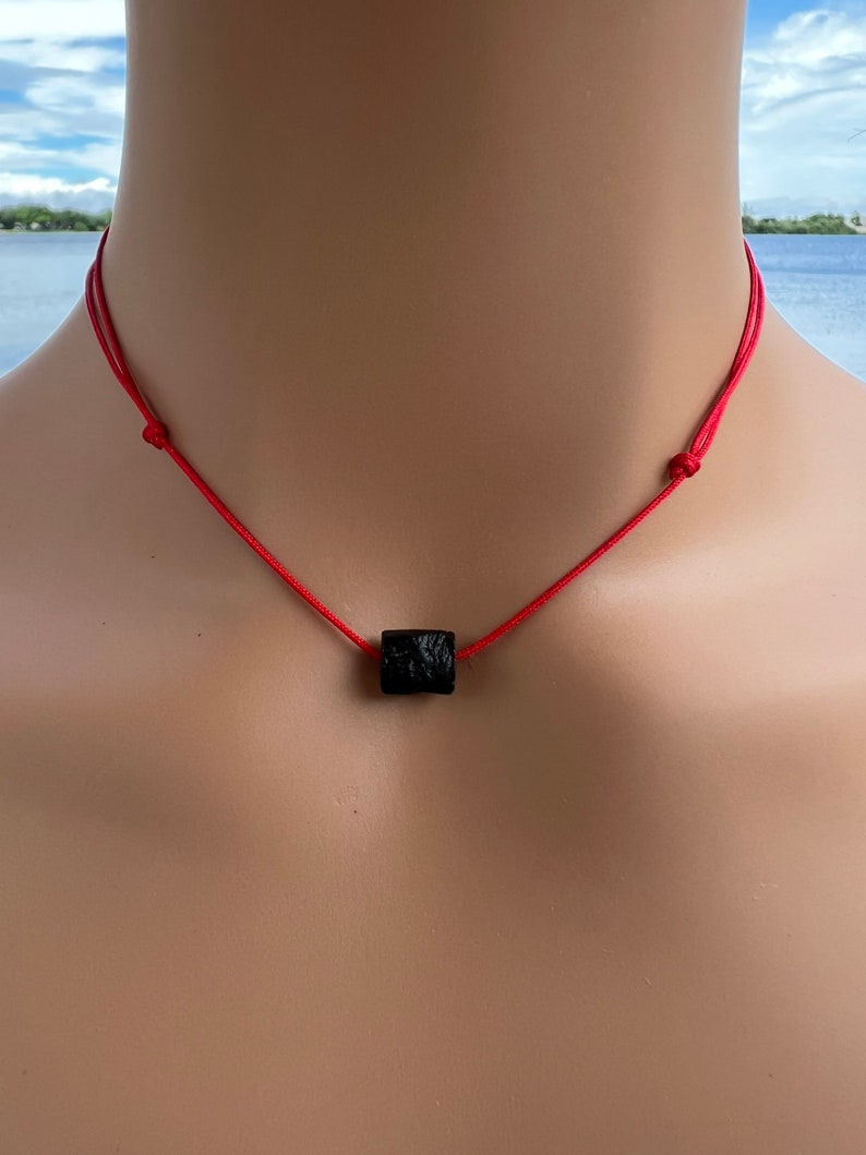 Puede incluir: Un collar de cuerda roja con un colgante cuadrado negro.
