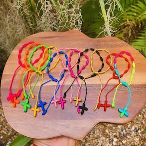 Puede incluir: Una colección de pulseras anudadas de colores con un dije de cruz en el extremo. Las pulseras están hechas de cuerda y vienen en una variedad de colores, incluyendo rojo, naranja, amarillo, verde, azul, morado, rosa y negro.