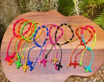 May include: A collection of colorful knotted bracelets with a cross charm at the end. The bracelets are made of string and come in a variety of colors, including red, orange, yellow, green, blue, purple, pink, and black.