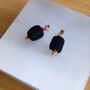 May include: Two black pendant charms with gold accents. The charms are shaped like a cylinder and have a small red bead on one end.