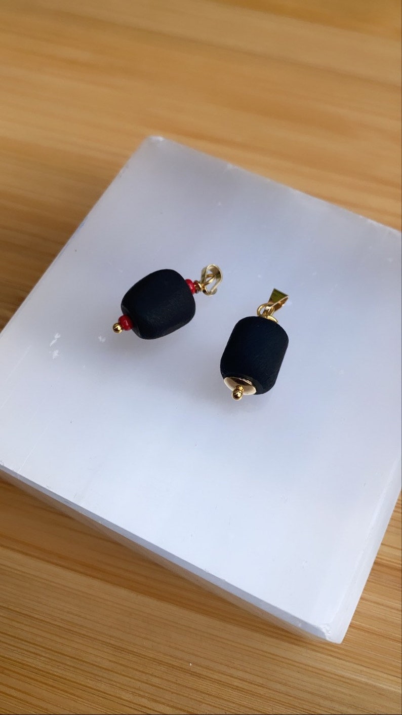 May include: Two black pendant charms with gold accents. The charms are shaped like a cylinder and have a small red bead on one end.