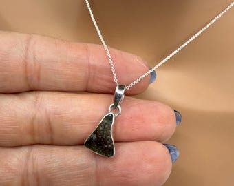 May include: A silver necklace featuring a pendant with a dark green, triangular stone. The stone has a textured surface and is suspended from a delicate silver chain. The pendant is held in place by a small silver bail.
