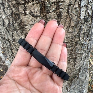 May include: A black rubber bracelet with a rectangular black metal charm. The charm has a faceted design.