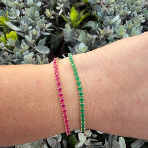 May include: Two delicate bracelets, one with vibrant pink gemstones and the other with vivid green gemstones, are displayed on a wrist. The bracelets are set in a gold-toned metal, creating a striking contrast against the skin and the green foliage in the background.