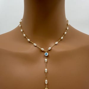May include: A gold-toned necklace featuring a strand of small, white pearls. The necklace has a drop pendant with a blue eye charm, more pearls, and a gold-colored Hamsa hand charm. The necklace is displayed on a person.