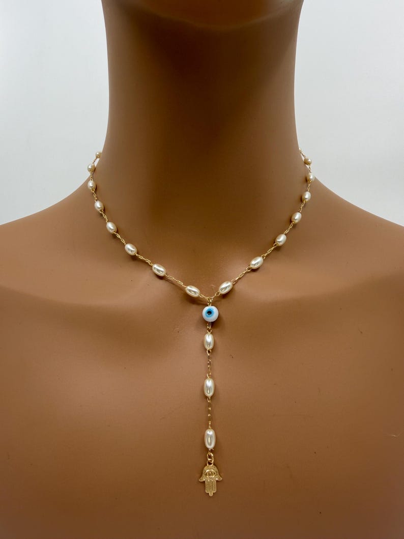 May include: A gold-toned necklace featuring a strand of small, white pearls. The necklace has a drop pendant with a blue eye charm, more pearls, and a gold-colored Hamsa hand charm. The necklace is displayed on a person.
