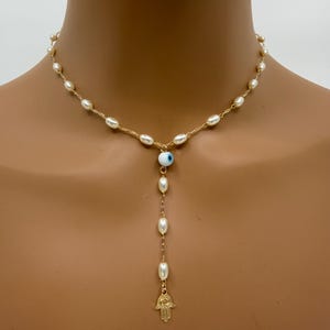 May include: A gold-toned necklace featuring a strand of small, off-white pearls. The necklace has a Y-shaped pendant with a blue and white evil eye charm, and a gold-toned Hamsa hand charm at the end of the pendant.