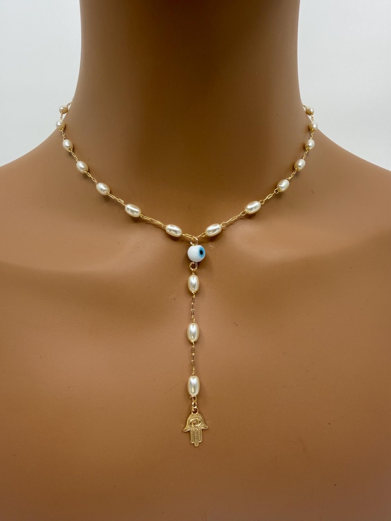 May include: A gold-toned necklace featuring a strand of small, off-white pearls. The necklace has a Y-shaped pendant with a blue and white evil eye charm, and a gold-toned Hamsa hand charm at the end of the pendant.
