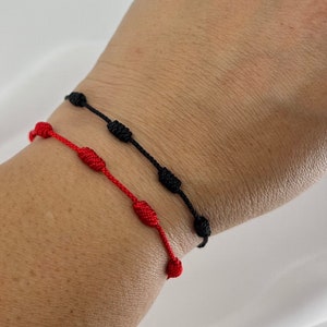 Op de afbeelding: Twee vriendschapsarmbanden, een rode en een zwarte, worden gedragen om de pols van een persoon. De armbanden zijn gemaakt van touw en hebben knopen op regelmatige afstanden.