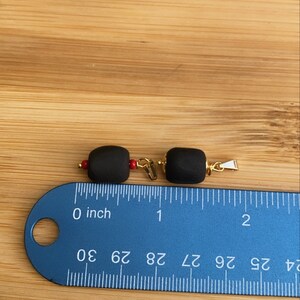 May include: Two black beads with gold accents and red accents. The beads are about 1 inch long and are measured on a ruler.
