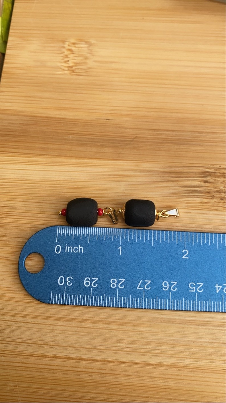 May include: Two black beads with gold accents and red accents. The beads are about 1 inch long and are measured on a ruler.