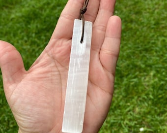 May include: A white selenite crystal pendant on a brown leather cord. The crystal is smooth and has a striated texture.