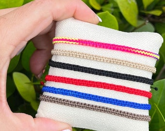 May include: A set of six colorful friendship bracelets on a white fabric background. The bracelets are made of woven string in various colors, including red, blue, black, beige, and pink.
