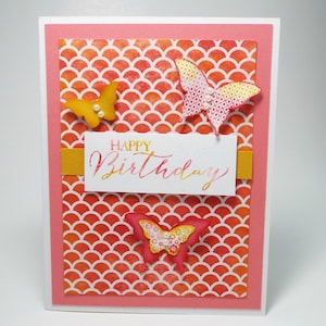 May include: A pink card with a white background and the words "Happy Birthday" written in gold script. The card is decorated with three paper butterflies in shades of orange, pink, and yellow. The butterflies have white accents and are layered on top of a red and orange patterned background.