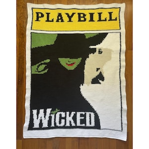 May include: A crocheted blanket featuring the words "PLAYBILL" in black on a yellow background. Below, the blanket depicts two characters from the musical "Wicked" in black, green, and white, with the word "WICKED" in white.