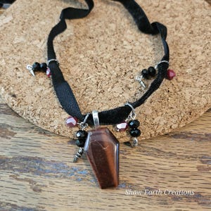 Op de afbeelding: Een zwarte fluwelen choker ketting met een doodskistvormige hanger van bruine tijgeroogsteen. De ketting heeft zwarte en rode kralen en kleine zilverkleurige bedels. De tekst "Shaw Earth Creations" is zichtbaar.
