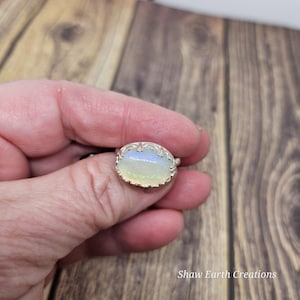 May include: Oval-shaped opal ring with a silver filigree setting. The opal has a milky white base with iridescent flashes of blue and yellow. The ring is held in a hand, showcasing the intricate details of the jewelry. The text "Shaw Earth Creations" is visible.