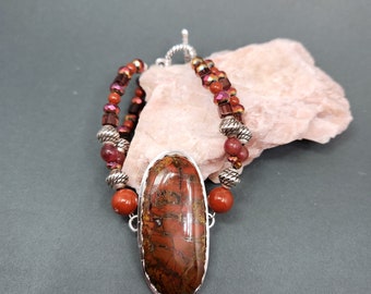 Op de afbeelding: Een ketting met een rode en bruine edelsteen hanger, zilveren kralen en een zilveren sluiting. De ketting ligt op een roze steen.