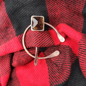 Puede incluir: Un broche de metal plateado y marrón con una piedra de forma cuadrada en el centro. El broche está sobre una tela de cuadros rojos y negros.
