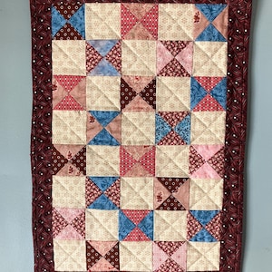 May include: A small, quilted wall hanging with a patchwork design. The quilt features squares of cream, pink, blue, and brown fabrics. A dark red border with white dots surrounds the quilt. It is hung from a wooden dowel.
