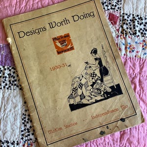 May include: Vintage "Designs Worth Doing" booklet from McKim Studios, 1930-31, featuring a black and white illustration of a woman quilting. The booklet is on a pink and patterned quilt.
