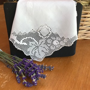 May include: A white handkerchief with a delicate lace trim, folded and placed on top of a black purse. A bouquet of lavender flowers is in the foreground.