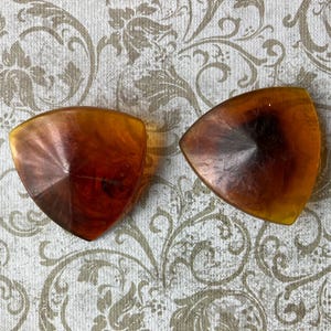 May include: Two vintage, amber-colored, triangular-shaped clip-on earrings. The earrings have a marbled appearance with a dark brown center and lighter amber edges. They are set against a patterned, off-white background.