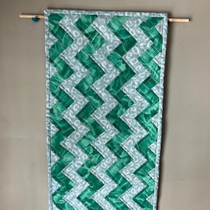 May include: A rectangular wall hanging with a green and white chevron pattern. The quilt is suspended from a wooden dowel rod with a blue bead. The background is a solid gray wall.