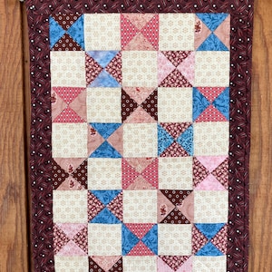 May include: A handmade quilt with a patchwork design. The quilt features squares of cream, pink, blue, and brown fabrics. The border is a dark burgundy with a small white pattern. The quilt is hanging on a wooden rod.