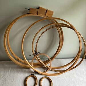May include: A collection of wooden embroidery hoops in various sizes, including several large circular hoops with metal tightening screws and two smaller oval-shaped hoops with textured inner rims, all arranged together in a group.