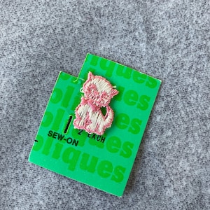 May include: A pink and white embroidered cat patch on a green tag. The tag reads "Sew-On" and "2 Each". The patch is a detailed design of a cat with its tail curled. The background is a gray fabric.
