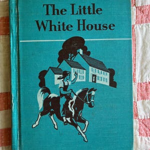 May include: A hardcover book titled The Little White House with a teal cover. The front features a dark brown illustration of a person riding a horse in front of a house with trees. The text is printed in dark brown lettering at the top.