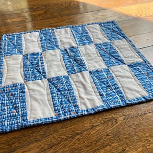 May include: A square, quilted placemat with a checkerboard pattern. Alternating squares are white and blue, with the blue squares featuring a repeating triangle design and small orange accents. The placemat is approximately 12 inches square.