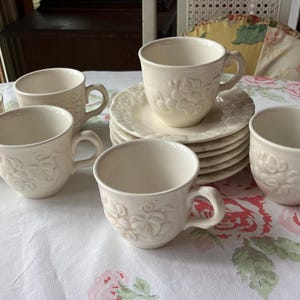 May include: A collection of off-white teacups and saucers arranged on a floral tablecloth. The cups and saucers have a raised floral design. The set includes six teacups and a stack of saucers. The teacups have handles.