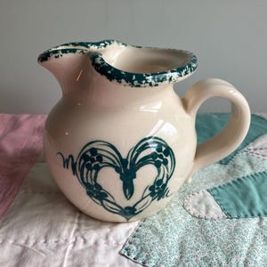 May include: Cream-colored ceramic pitcher with a heart-shaped design in teal. The pitcher has a curved handle and a scalloped rim with teal detailing. The heart design is composed of floral elements. A decorative kitchenware item.