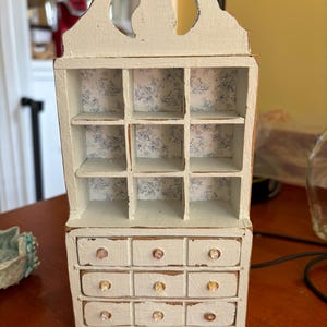 May include: A white painted miniature hutch with a distressed finish. The hutch has a top shelf with nine square compartments and a bottom section with three drawers. Each drawer has three crystal-like knobs.