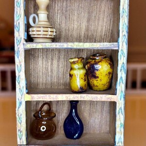 May include: A miniature wooden shelf unit with three shelves, decorated with a black and white checkered pattern and floral designs. The shelves are filled with small decorative items, including a brown and white pitcher, two yellow and brown ceramic vases, a brown glass bottle, and a blue glass bottle. The shelf unit has a quote on the front that reads "He had an adventure Par..."