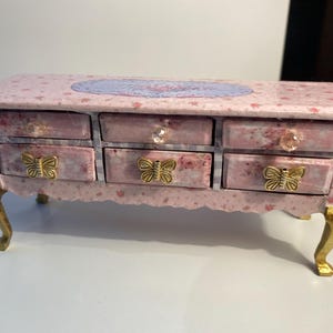 May include: A pink wooden dresser with six drawers and gold butterfly drawer pulls. The dresser has a floral pattern and a light blue oval in the center of the top.