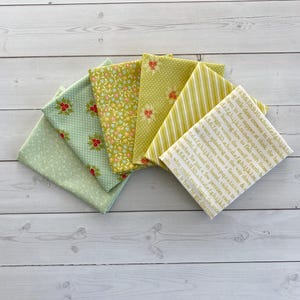 May include: A collection of folded fabric squares in shades of green and yellow, featuring various patterns like floral, polka dots, and stripes. One piece has text printed on it. Ideal for quilting and crafting projects.