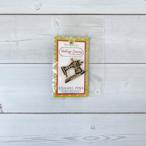 May include: A gold enamel pin featuring a vintage sewing machine with floral details. The pin is packaged on a card with the text "Fig Tree & Co Vintage Sewing Collection" and "XOXOXO".