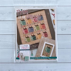 May include: A cross-stitch pattern kit titled Prim Village featuring a grid of colorful houses on a tan background, accompanied by a small enamel keychain charm shaped like a house, all packaged in a clear plastic sleeve with branding text.