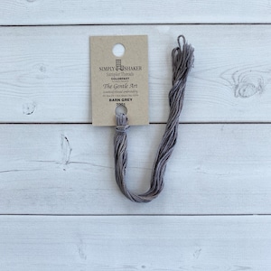 May include: A skein of grey embroidery thread attached to a tan cardboard tag. The tag features the text Simply Shaker Sampler Threads, The Gentle Art, counted thread embroidery, and the color name Barn Grey 7001 with a small chair icon.