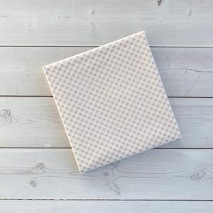 May include: A folded square of fabric with a repeating pattern of small, light brown squares on a white background. The fabric is resting on a light-colored wooden surface.