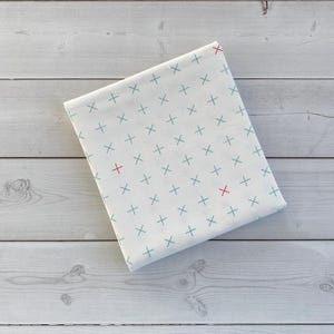 May include: Folded white fabric with a repeating pattern of teal and red plus signs. The fabric is resting on a light gray wooden surface. The plus signs are evenly spaced across the fabric.