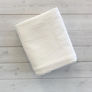 May include: Folded white fabric with a pattern of small, evenly spaced grey dots. The fabric is soft and appears to be cotton or a similar material. The background is a light-coloured wooden surface.