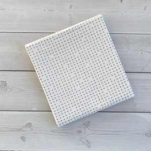May include: Folded white fabric with a repeating gray cross pattern. The fabric is neatly folded and rests on a light gray wooden surface. The pattern is evenly spaced and covers the entire surface of the fabric.