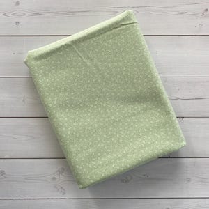 May include: A folded bundle of fabric in a soft green hue, adorned with a delicate, all-over floral pattern in a lighter shade of green. The fabric rests on a light-colored wooden surface.
