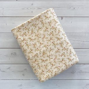 May include: A folded fabric featuring a cream-coloured base with a repeating floral design. The pattern incorporates small, brown and red flowers and green leaves. The fabric rests on a light grey wooden surface.